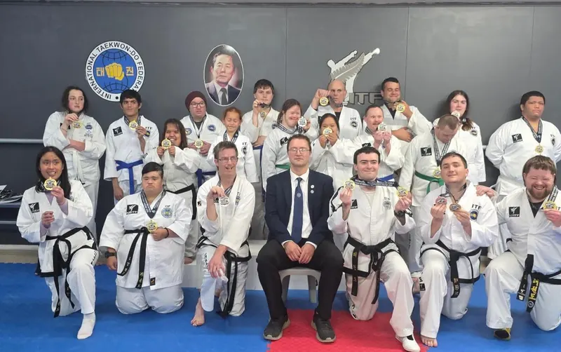 A large group of Inclusive Taekwon-Do athletes holding up their medals in the dojang, with Ben Evans seated in the centre